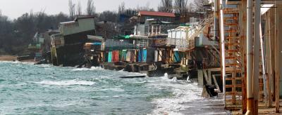 Beachfront buildings extensively damaged by erosion with portions collapsed into the sea, and waves lapping against the remains of the structures. Trees are visible in the background.