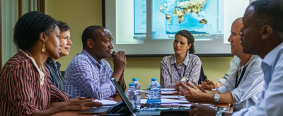 Six people sit around a conference table in a meeting room, with a world map displayed on a projection screen behind them.