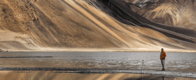 A person stands near a tripod by a reflective mountain lake with snow-covered peaks in the background.