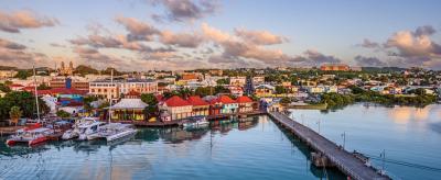 Coastal cityscape with colorful buildings along the waterfront, boats docked at a pier, and a calm sea reflecting the blue sky and scattered clouds.