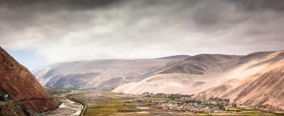 A scenic view of a river winding through agricultural fields and a small settlement, surrounded by rolling hills under a cloudy sky.