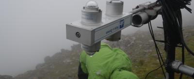 A person in a green jacket adjusts equipment on a foggy, rocky hilltop.