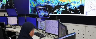 A person in a hijab works at a desk with multiple monitors displaying weather data and maps in a control room. Large screens on the wall show satellite images.