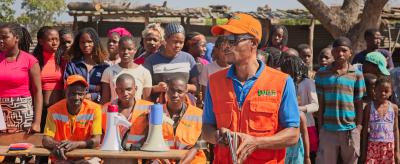 A group of people, including several in orange vests, stand outdoors near tables with megaphones. One person in an orange cap addresses the crowd.