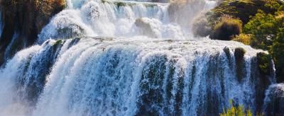 Multilevel waterfall cascading over rocky terrain surrounded by lush greenery under a clear sky.