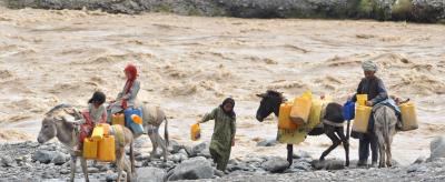 People and donkeys carrying yellow containers navigate a rocky riverbank with a fast-flowing river in the background.