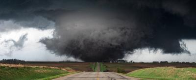 A large, dark tornado approaches a rural road, flanked by open fields under an ominous sky.