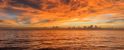 Sunset over the ocean with vibrant orange and yellow hues in the sky, reflecting on the water's surface. Waves gently lap against the shore under dramatic cloud formations.