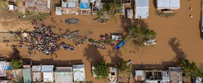 Aerial view of a flooded village with submerged buildings and boats. A large group of people gathers on dry ground, surrounded by muddy water. Trees and damaged structures are visible.