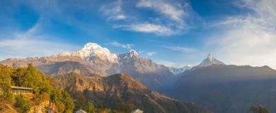 A Himalayan village with traditional houses sits in the foreground, surrounded by green hills. Snow-capped mountains rise under a clear blue sky in the background.