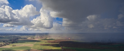Aerial view of patchwork farmland under partly cloudy skies, with sunlight on one side and rain clouds casting shadows on the other.