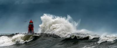 A red lighthouse stands amid turbulent waves crashing against a rocky shore under a dark, cloudy sky.