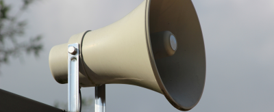 A close-up of a gray outdoor loudspeaker mounted on a metal bracket against a cloudy sky background.