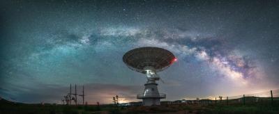 A large radio telescope dish stands under a star-filled night sky with the Milky Way galaxy visible overhead.