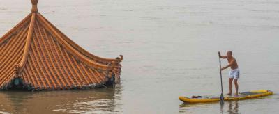 A person on a paddleboard navigates through floodwaters near a partially submerged traditional gabled roof.
