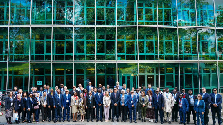 A large group of individuals in formal attire stands in several rows in front of a modern glass building, posing for a group photo.