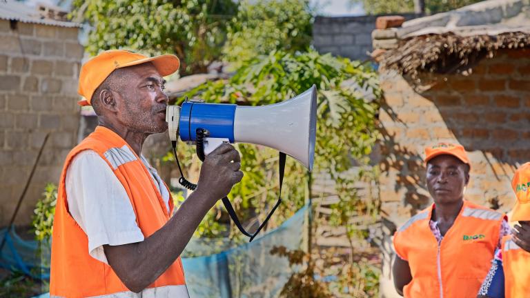 Man in an orange vest and cap speaks into a megaphone outdoors. Another person in similar attire stands nearby.