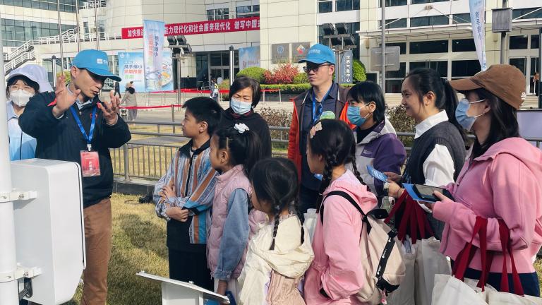 A group of adults and children, some wearing masks, listen attentively to a person speaking outside a modern building. The scene suggests an educational or guided tour.