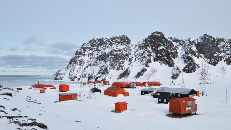 A cluster of red and black buildings sits on snow-covered ground near rocky, snow-capped mountains and the sea under a cloudy sky.