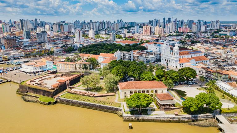 Aerial view of a historic riverside district with colonial buildings, a fort, and modern city skyline in the background under a partly cloudy sky.