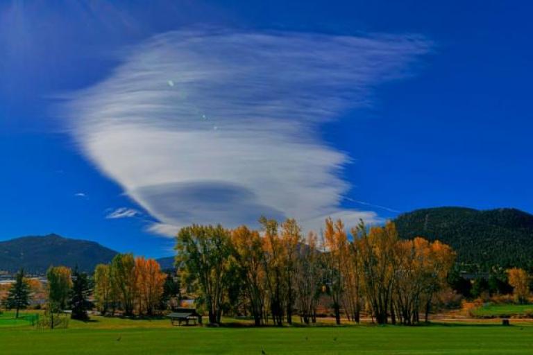 A cloud over a field with trees and mountains in the background.