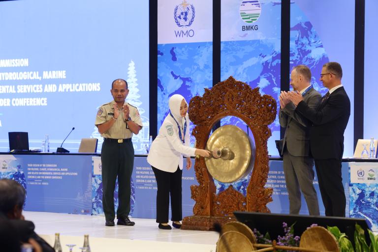 A woman strikes a ceremonial gong, while 3 other people applaud.