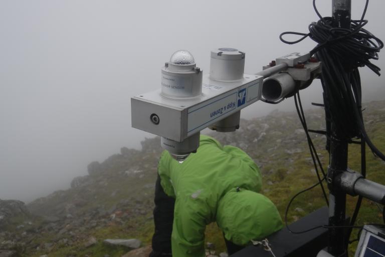 A person in a green jacket adjusts scientific monitoring equipment in a foggy, mountainous setting.