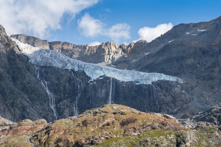 Glacier with ice cascading down between rocky peaks under a clear blue sky, flanked by waterfalls and rugged terrain.