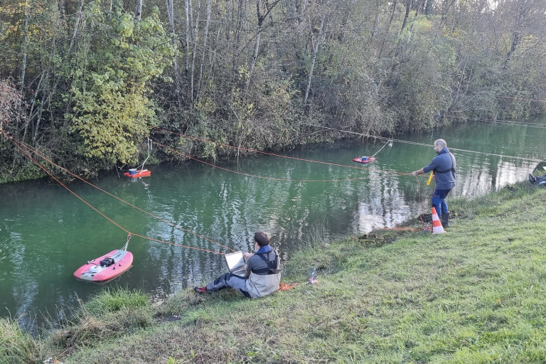 Two people on a riverside using remote-controlled boats, one seated with a laptop and the other standing, in a lush green setting.