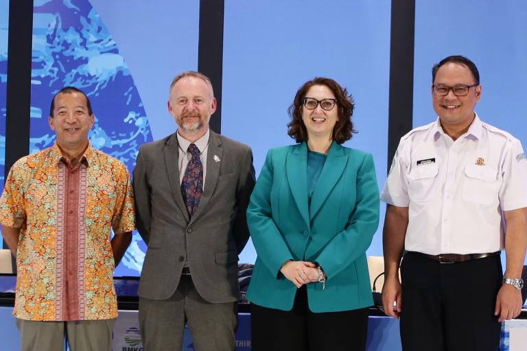 Four individuals standing side by side in formal attire at a conference or event with a world map in the background.