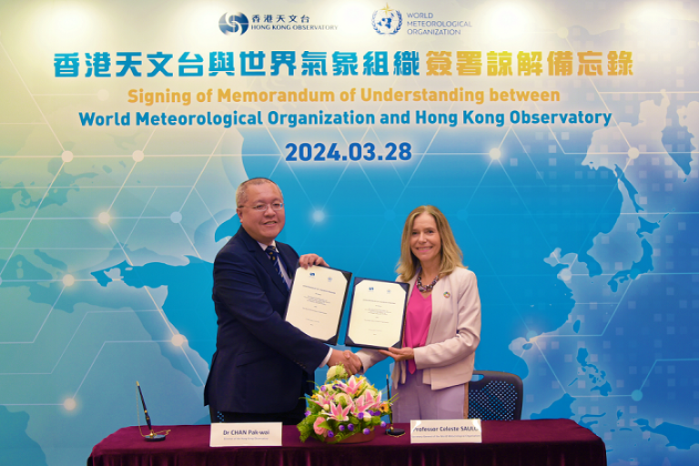 Two individuals exchanging a signed memorandum at a ceremony with a backdrop displaying logos of the world meteorological organization and hong kong observatory.