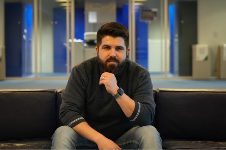 A man with a beard sits on a dark sofa in an office space, resting his chin on his hand. He wears a dark sweater, jeans, and a smartwatch. The background features glass doors and office decor.