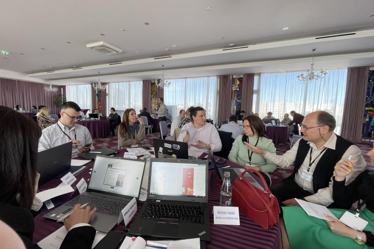 Professionals in a business meeting at a conference table, engaging in discussions with laptops open in a well-lit room with large windows.