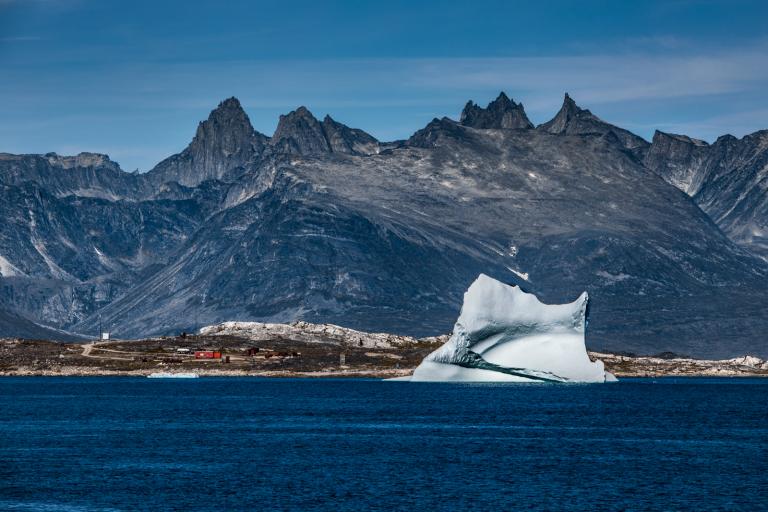 A large iceberg floats in a calm blue sea with rocky mountains in the background under a clear sky.