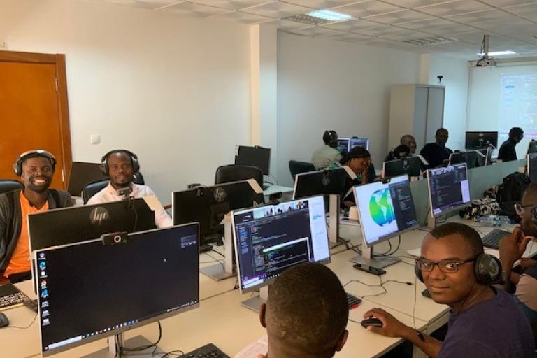 A room with multiple people wearing headphones and working on computers. Some screens show coding or data visualization programs. The environment appears to be collaborative and focused.