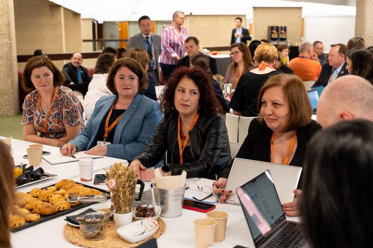 A group of people sit around a table with laptops and pastries, engaged in discussion at a conference or meeting.
