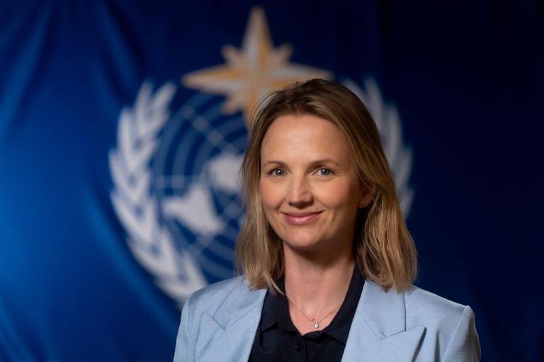A person with shoulder-length blonde hair wearing a light blue blazer smiles in front of a blurred background featuring a blue flag with a white emblem.