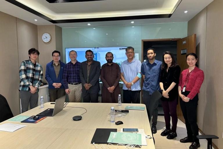A group of eight people standing and posing for a photo in a conference room with a table, a laptop, and documents.