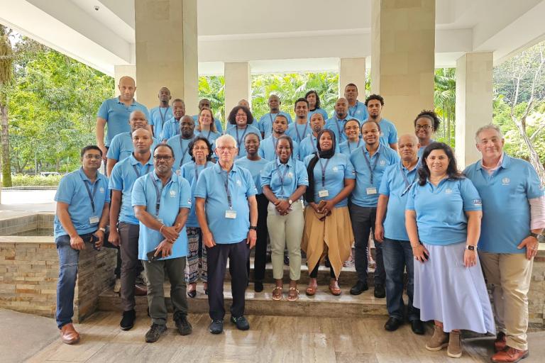 A group of 31 people wearing light blue polo shirts and name badges pose for a photo in a spacious indoor area with columns and greenery visible in the background.