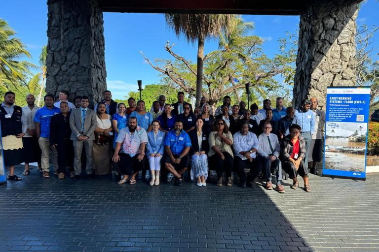 A group of people stand together outside on a paved area under a wooden structure, with trees and a blue sky in the background. A large informational poster is displayed on the right side of the group.