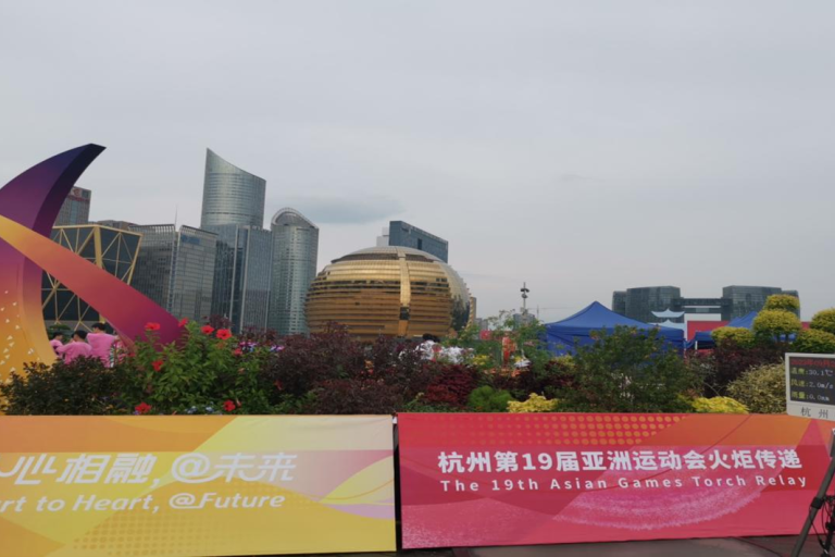 The image shows a public display with banners and signage for the 19th Asian Games Torch Relay in Hangzhou, including the slogan "Heart to Heart, @ Future," set against urban buildings and greenery.