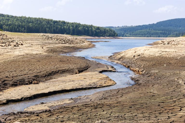 A narrow stream flows through parched, cracked earth with forests and hills in the background under a partly cloudy sky.