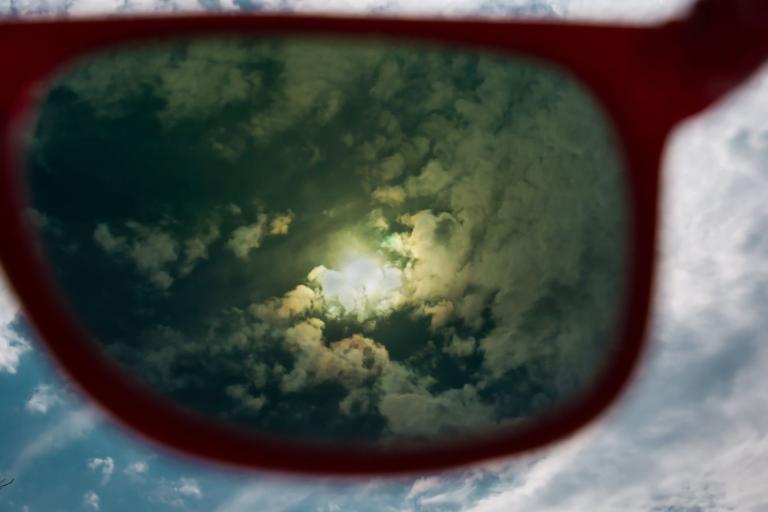 View of cloudy sky with a bright sun seen through red-framed sunglasses.