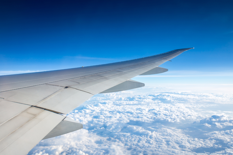 View of an airplane wing flying over a layer of clouds with a clear blue sky in the background.