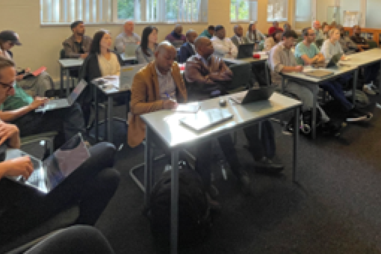 A group of people seated in a classroom, each with laptops or notepads, attentively listening to a presentation.
