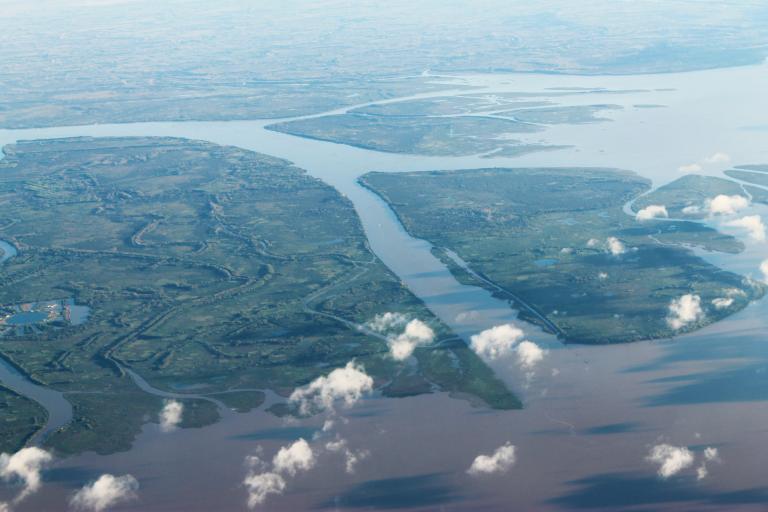 Aerial view of a river delta with multiple branching waterways and green landmasses, partially obscured by scattered clouds.