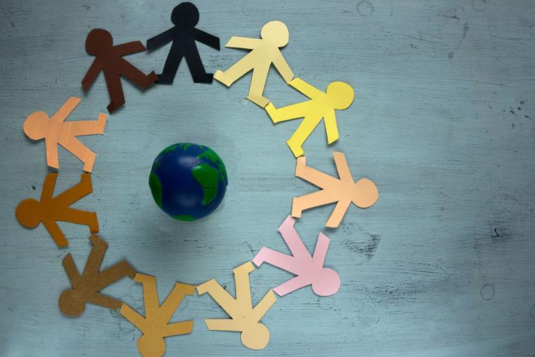 A ring of multicolored paper cutouts of people holding hands encircles a small globe on a gray background.