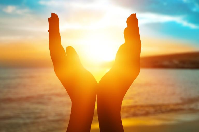Hands are cupped together in front of a sunset over a beach, framing the sun. The sky has vibrant shades of orange, yellow, and blue.