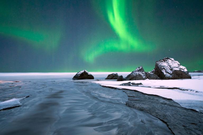 A stunning display of the Northern Lights illuminates the night sky over a snowy landscape with scattered rocks and a frozen body of water.