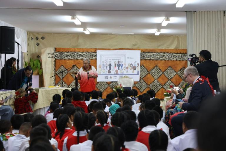 A speaker addresses a group of schoolchildren seated on the floor in a decorated room with a projected presentation behind them. Several adults are standing, and a cameraman is filming the event.